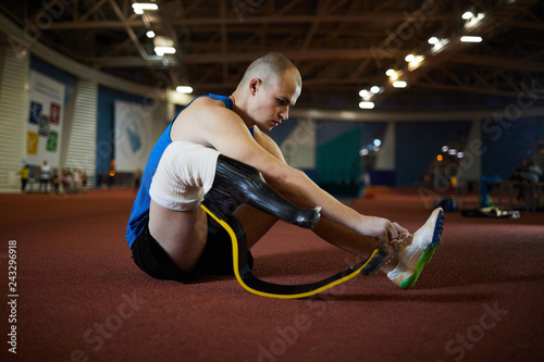 Active man with artificial right leg tying shoelace while sitting on the floor of modern stadium