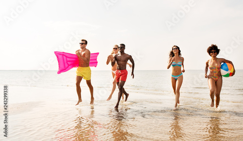friendship, summer holidays and swimming concept - happy friends with floating mattress and inflatable ball running on beach