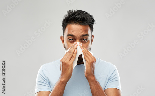 health problem, rhinitis and allergy concept - unhealthy indian man with paper napkin blowing nose over grey background