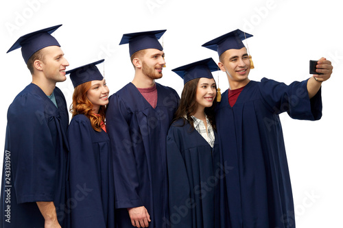 education, graduation and people concept - group of happy graduate students in mortar boards and bachelor gowns taking selfie by smartphone over white background