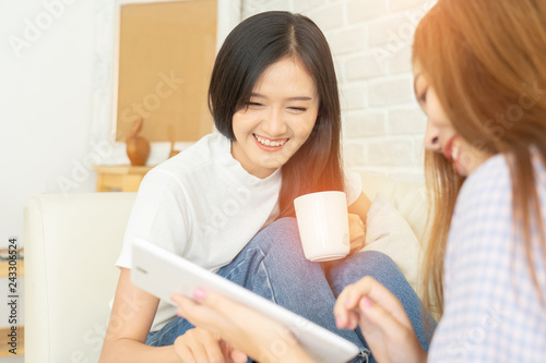 Happy female friends holding coffee mugs while discussing at table in living room relax time.Asian beauty woman smile and sititing sofa .she shopping online .