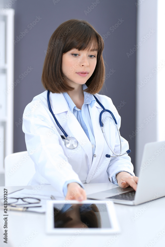 Doctor typing on laptop computer while sitting at the white table in hospital office. Physician at work. Medicine and healthcare concept Doctor typing on laptop computer while sitting at the white table in hospital office. Physician at work. Medicine and healthcare concept