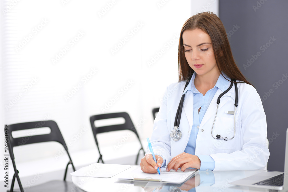 Doctor woman at work. Portrait of female physician filling up medical form while sitting at the glass desk at clinic or hospital. Medicine and healthcare concept