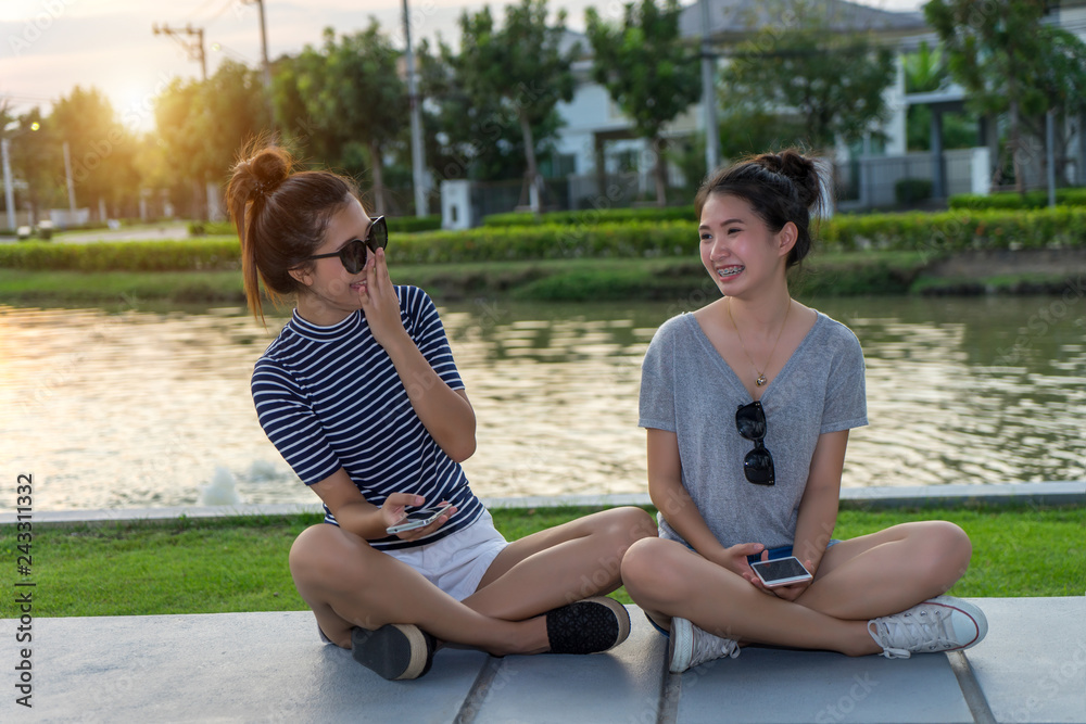 Happy women friends talking and laughing gossip holding mobile phone in a park outdoors with green background. Two young Asian girl with friends time sitting outside joyful having fun together.