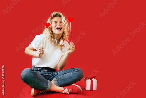 beautiful young girl with red hearts and a white gift with a bow sits in full growth on an isolated red backgroundValentine's Day. photo