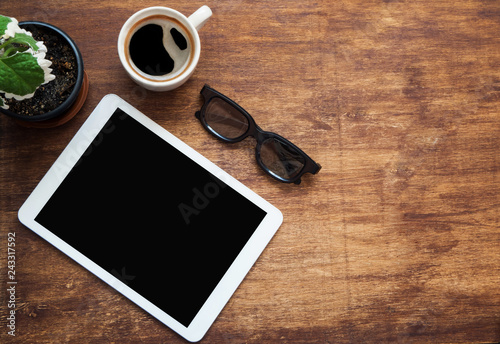 Tablet blank screen, black glasses cup of coffee and green flower on wood desk. Copy space. Flat lay