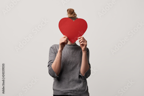 woman hiding face behind blank paper cut heart card isolated on grey