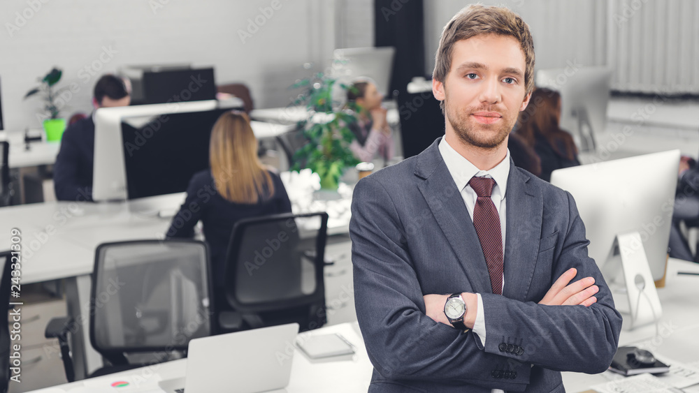 handsome confident young businessman standing with crossed arms and looking at camera in office handsome confident young businessman standing with crossed arms and looking at camera in office