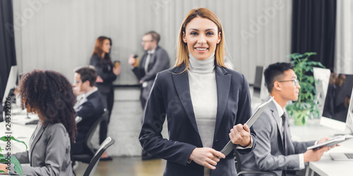 beautiful professional young businesswoman holding folder and smiling at camera in open space office