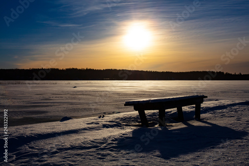 Winter landscape picture in Finland. Empty park bench in front. A frozen lake in the background. © Jarkko