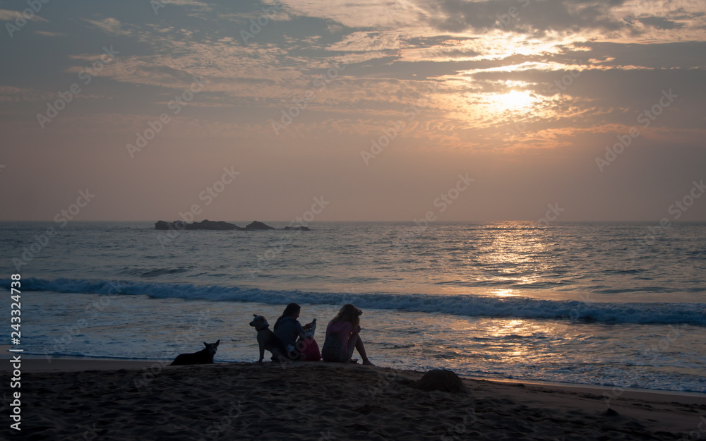 Two young women sit on the sandy beach of the ocean at sunset with gadgets