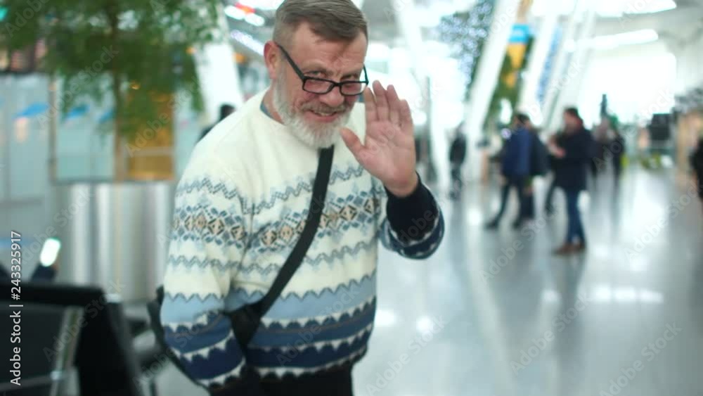 Mature man with a gray beard in a knitted sweater and glasses at the airport or train station. The pensioner waves goodbye and leaves carrying a suitcase on wheels behind him Mature man with a gray beard in a knitted sweater and glasses at the airport or train station. The pensioner waves goodbye and leaves carrying a suitcase on wheels behind him