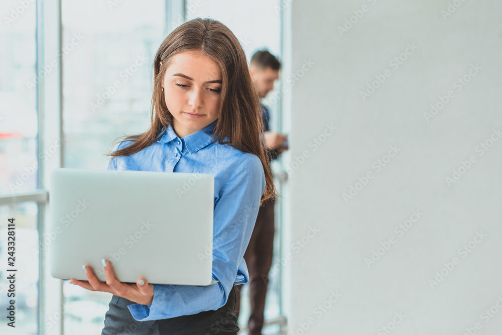 The secretary is standing in the office holding the laptop in his hands. During this she is on the background of her colleagues. The secretary is standing in the office holding the laptop in his hands. During this she is on the background of her colleagues.