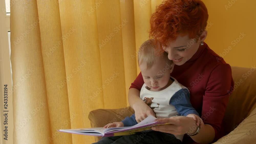 Mother with baby sitting in chair and reading a story with pictures. Little boy looks into the book and shows finger in book. Family, early development, activity, learning. Cute kid play with mom. Mother with baby sitting in chair and reading a story with pictures. Little boy looks into the book and shows finger in book. Family, early development, activity, learning. Cute kid play with mom.