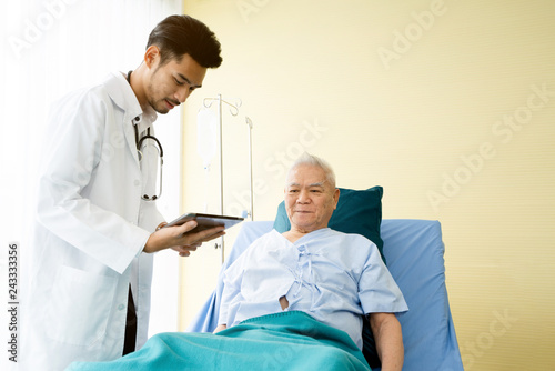 Asian Doctor is standing by explain the treatment information via the tablet to an elderly patient lying in bed in private sickroom. The treatment program is suitable for people to socialize elderly.