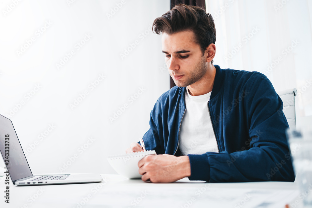 Attractive man working on laptop at space office while sitting table. Businessman writing in notepad work report Attractive man working on laptop at space office while sitting table. Businessman writing in notepad work report