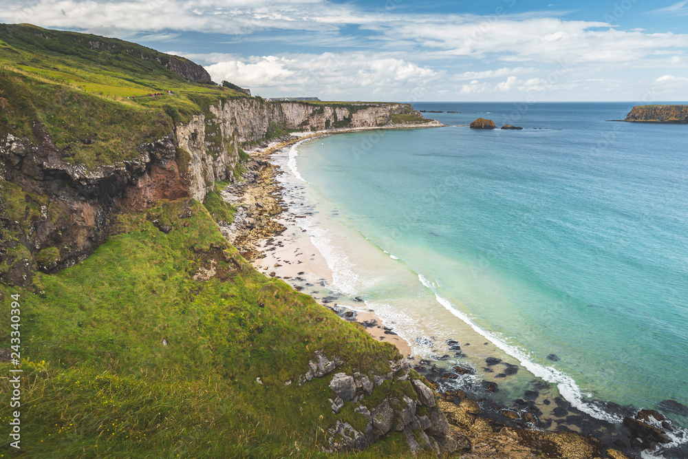 White sand beach of the Northern Ireland shoreline. Green grass covered cliff washed by the turquoise transparent sea water. Beauty of wild untouched environment. Breathtaking Irish landscape.