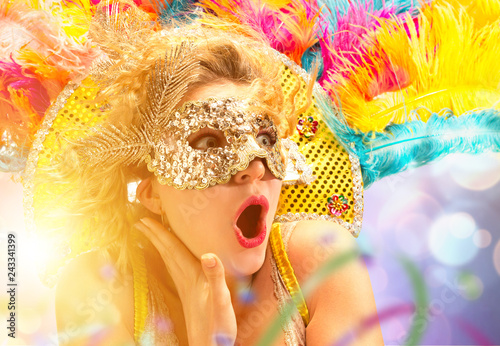 Beautiful surprised woman in carnival mask. Beauty model woman wearing masquerade mask at party over holiday background with magic glow. Christmas and New Year celebration. Glamour lady with perfect