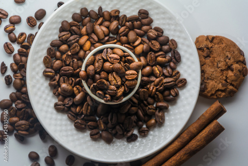 White Coffee cup with roasted coffee beans, cinnamon and biscuits on isolated white background from above. Coffee beans on plate and in the mug. Cinnamon and cookies on a table with coffee beans.