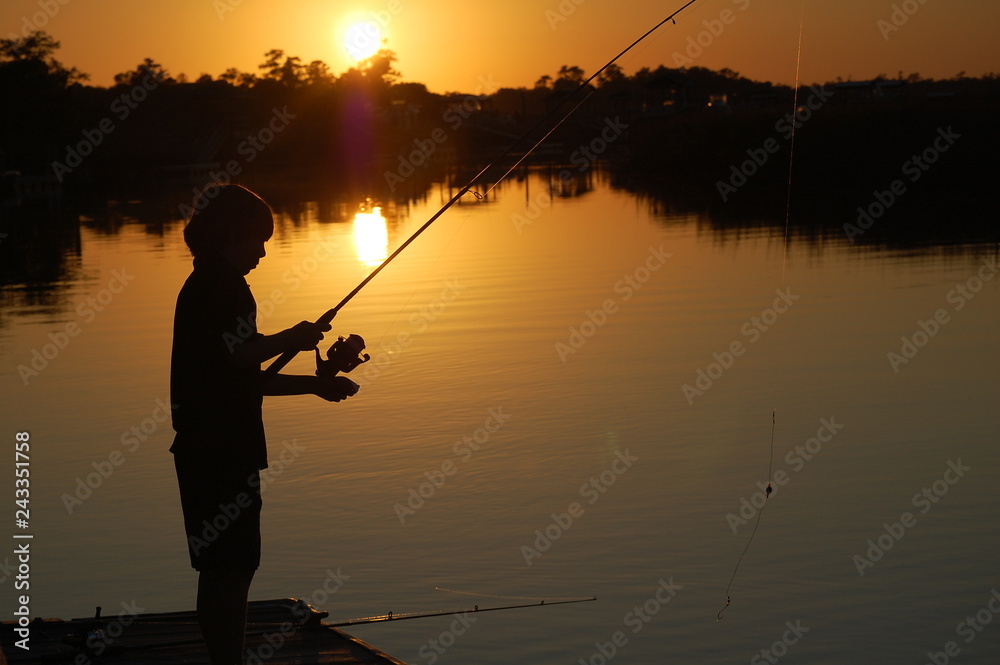 Silhouette of boy fishing off a dock at sunset. Stock Photo