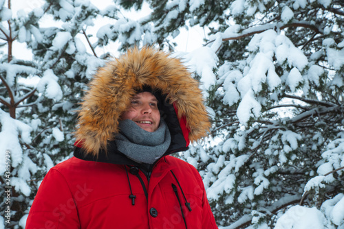 young adult man in red winter coat with hood with fur. snowed fir-tree on background