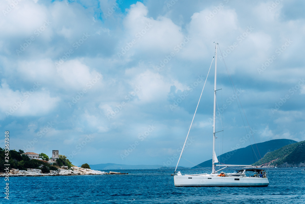 Summer scenery of port Fiskardo. White sail boat in beautiful tranquil bay opposite rocky seashore. Picturesque outdoor scene of Kefalonia island, Greece, Europe. Traveling vacation concept Summer scenery of port Fiskardo. White sail boat in beautiful tranquil bay opposite rocky seashore. Picturesque outdoor scene of Kefalonia island, Greece, Europe. Traveling vacation concept