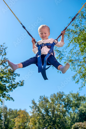 Emotional portrait of the little boy with seat belts on an attraction "Trampoline". He with delight soars in air against the background of the blue sky