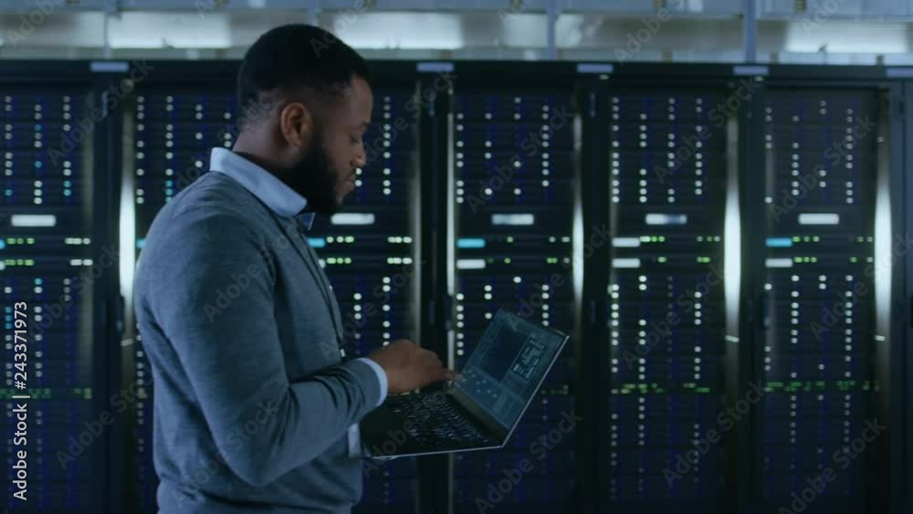 Black Data Center IT Technician Walking Through Server Rack Corridor with a Laptop Computer