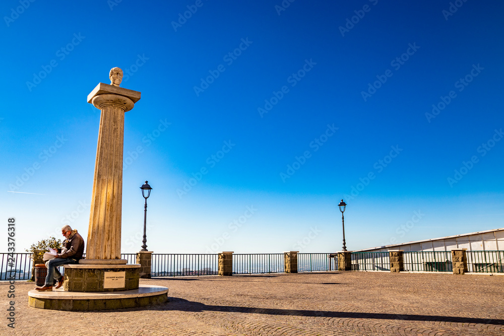 Travertine column with sculpture in memory of Giuseppe Mazzini, in the homonymous square with panoramic terrace. An old man reads a book. Ariccia, Castelli Romani, Lazio, Italy.