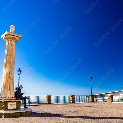 Travertine column with sculpture in memory of Giuseppe Mazzini, in the homonymous square with panoramic terrace. A boy looks at the smartphone. Ariccia, Castelli Romani, Lazio, Italy. photo