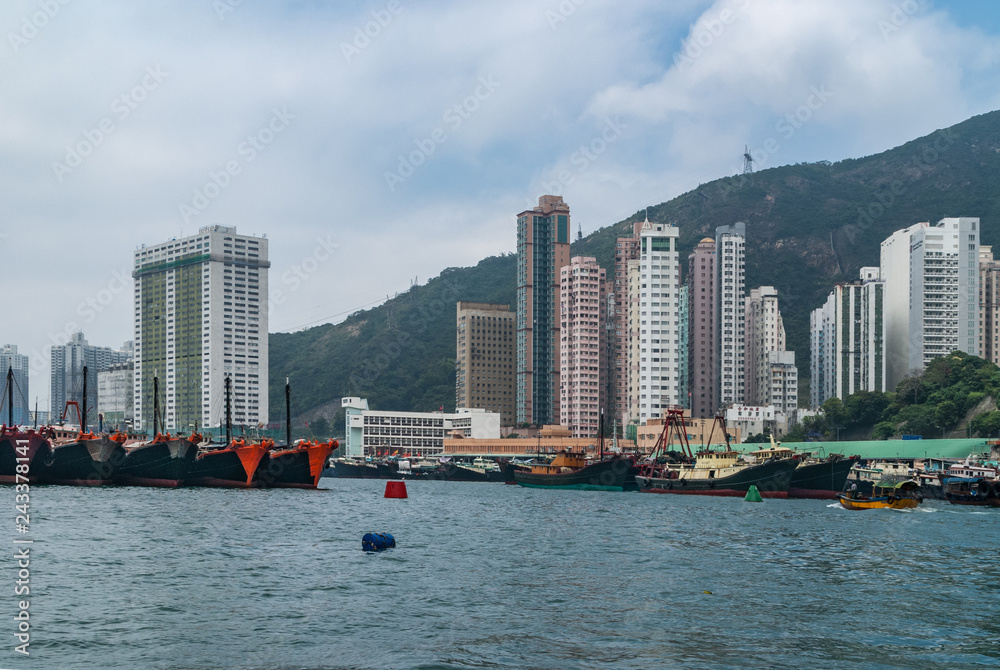 Fototapeta premium Hong Kong, China - May 12, 2010: Wide shot. Plenty black and red fishing vessels docked in harbor with many tall apartment buildings on shore. Clouded sky and green hill in back.