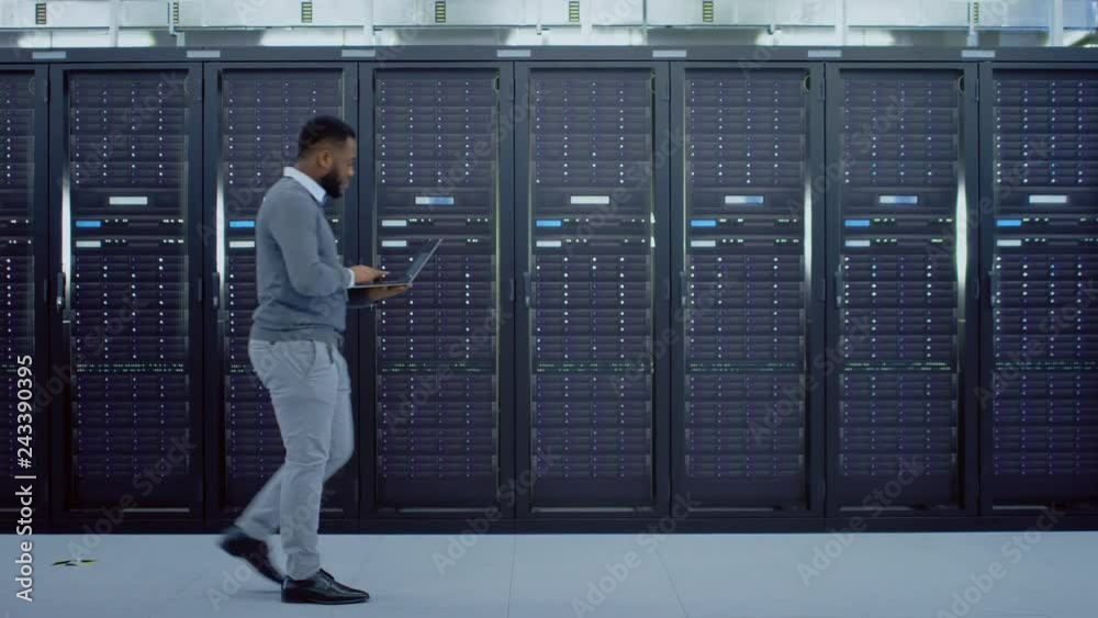 Black Data Center IT Technician Walking Through Server Rack Corridor with a Laptop Computer. He is Visually Inspecting Working Server Cabinets.