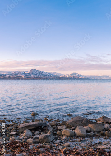 View of rocky shoreline, lake, and snow covered mountains at sunset
