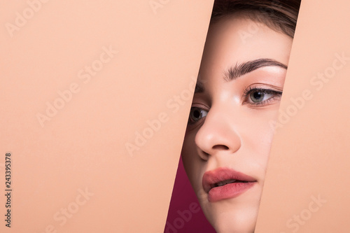 The face of a young beautiful girl with a bright make-up and puffy orange lips peers into a hole in peach paper.