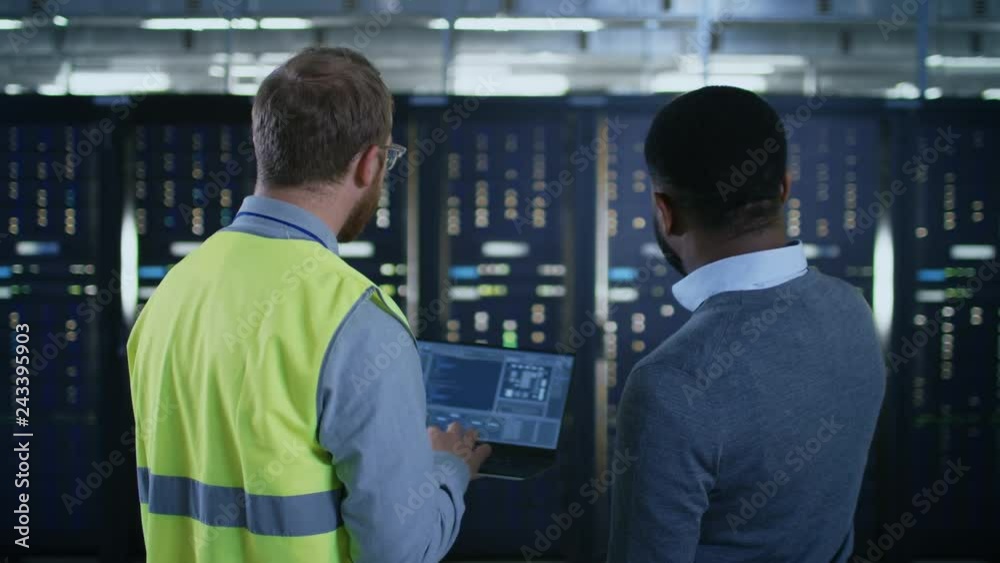 Bearded IT Engineer in Glasses and High Visibility Vest with a Laptop Computer and Black Technician Colleague Talking in Data Center while Standing Next to Server Racks. Run Diagnostics, Maintenance. Bearded IT Engineer in Glasses and High Visibility Vest with a Laptop Computer and Black Technician Colleague Talking in Data Center while Standing Next to Server Racks. Run Diagnostics, Maintenance.
