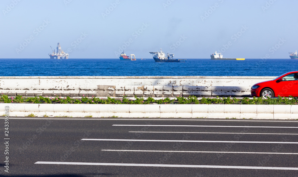 Red car heading to Las Palmas capital on coast road with oil platforms, offshore rig and big boats on sea on the background. Fossil fuel resources, environmental pollution concepts