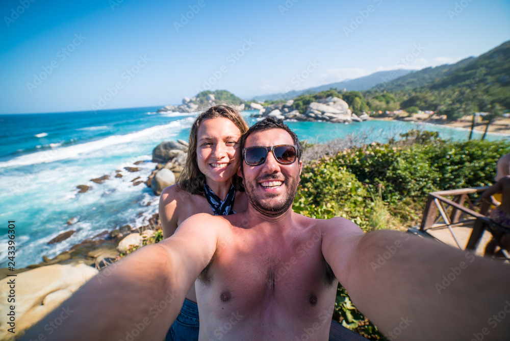 Obraz premium happy smiling couple taking selfie photo in front of the sea in Tayrona National Park, Tropical Colombia. Crazy tourists travelling on the white beach of jungles on caribbean sea.
