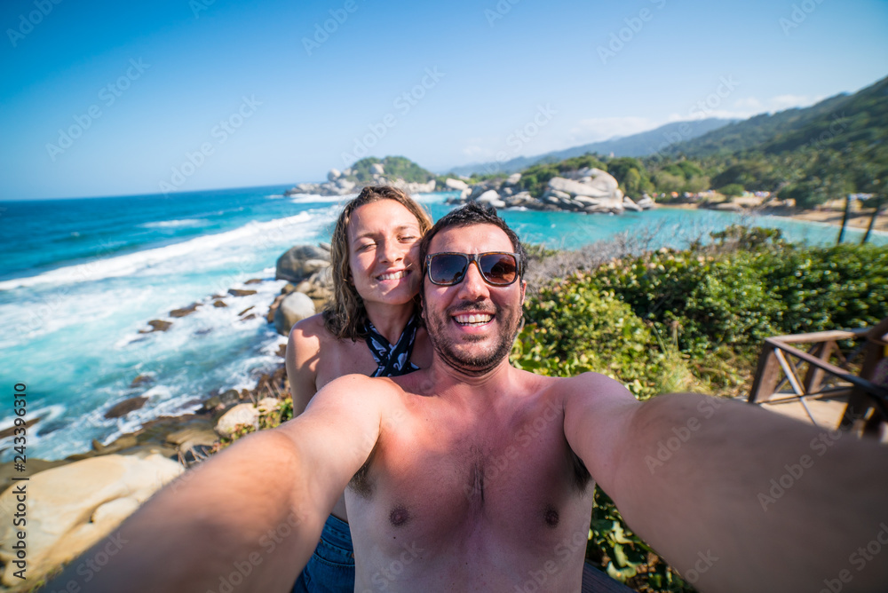 Obraz premium happy smiling couple taking selfie photo in front of the sea in Tayrona National Park, Tropical Colombia. Crazy tourists travelling on the white beach of jungles on caribbean sea. 