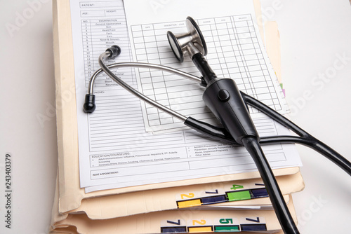 A stack of color-coded and numbered folders containing patient's medical records with blank doctor's visit form, ledger card, and stethoscope set on white background in horizontal image format.