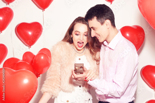 Man makes present to his lovely sweetheart girl. Lover's valentine day. Valentine Couple. Boy gives to his girlfriend jewelry. Wedding ring. Propose background red balloons hearts. Happy smile girl.