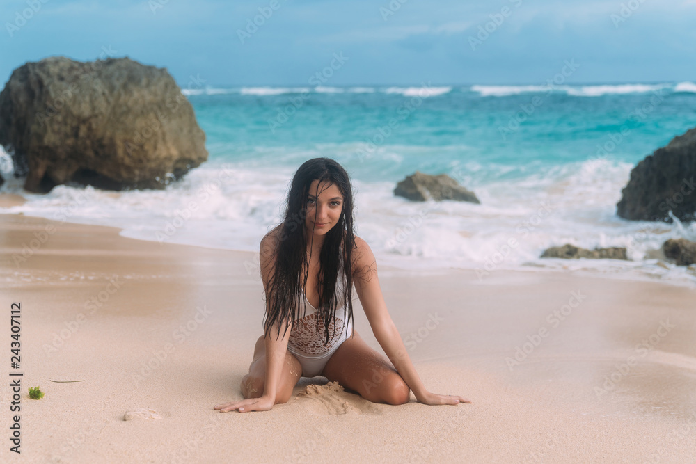 Sexy girl in swimsuit posing and sitting on deserted beach near ocean