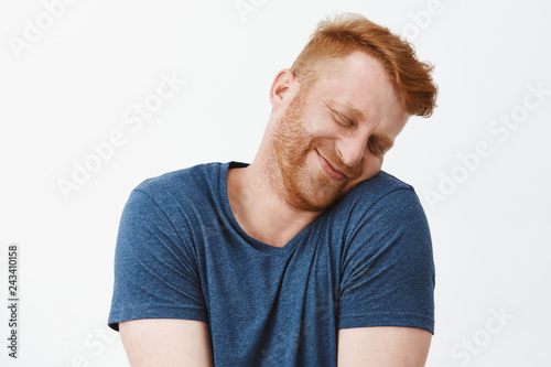 Silly and funny caucasian redhead guy with bristle, tilting head and leaning on shoulder with lovely and cute expression, smiling with closed eyes being tender and girly over gray background