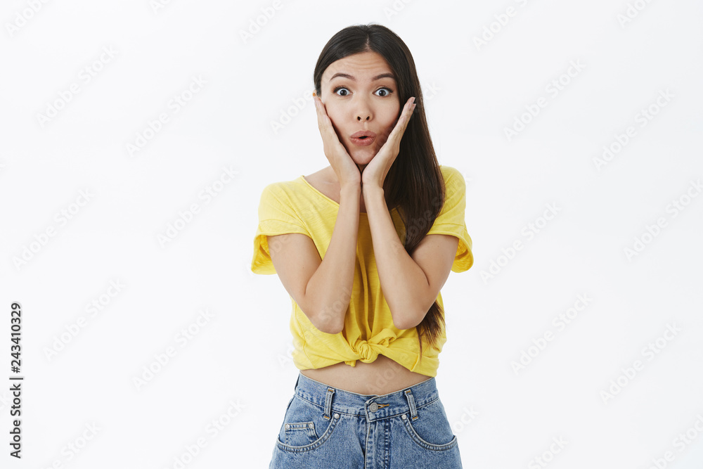 Portrait of enthusiastic excited and intrigued charming female shopoholic in yellow trendy t-shirt folding lips from amazement and surprised holding palms on cheeks posing astonished over white wall Portrait of enthusiastic excited and intrigued charming female shopoholic in yellow trendy t-shirt folding lips from amazement and surprised holding palms on cheeks posing astonished over white wall
