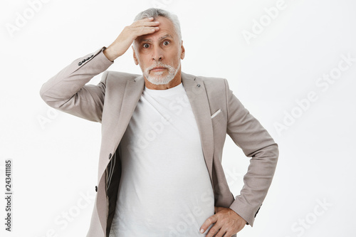 Portrait of nervous senior businessman in suit touching forehead holding hand on waist and holding breath feeling troubled and concerned facing problem standing worried and anxious over white wall