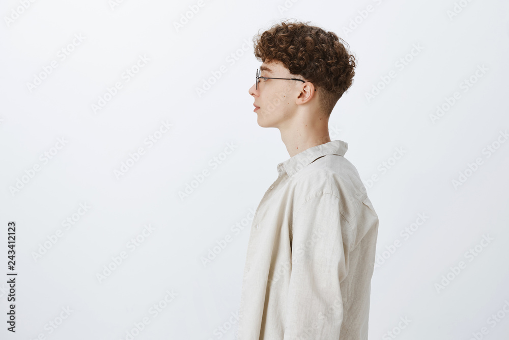 Obraz premium Profile shot of stylish handsome young hipster male freelancer with curly hairstyle in glasses looking left and standing casually over gray background in shirt, having no emotions as feeling calm