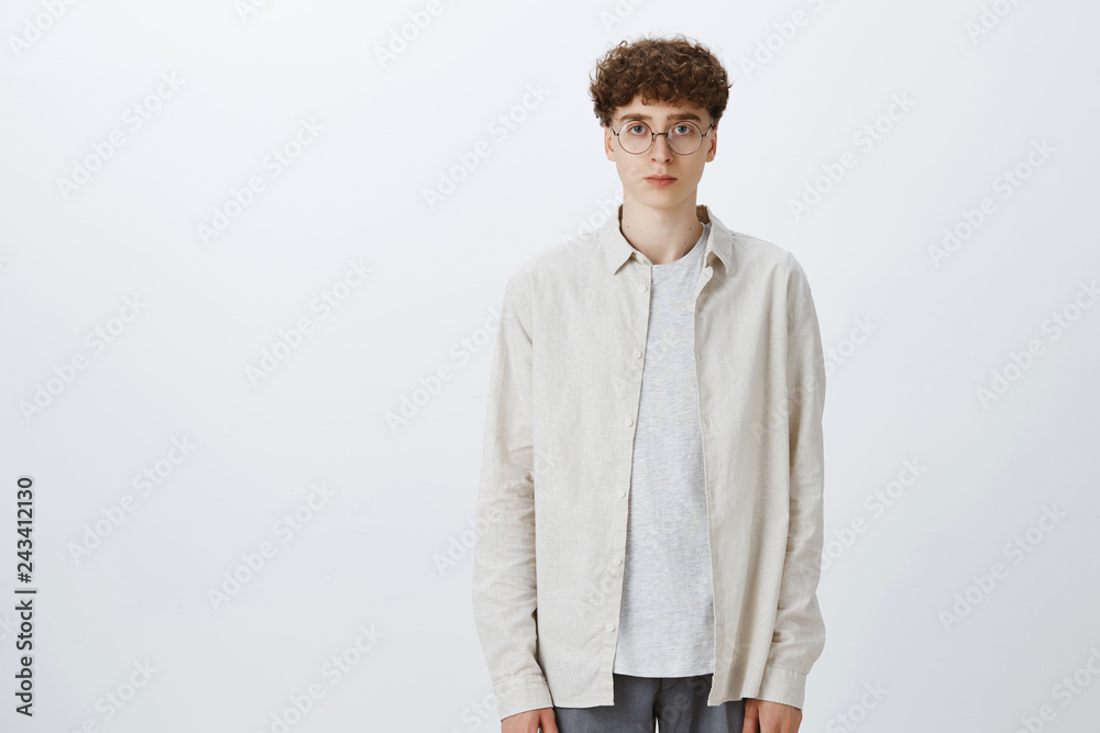 Fototapeta premium Studio shot of indifferent unemotional handsome and stylish european hipster boy in glasses with curly haircut standing upright looking at camera with no emotions and feelings, being calm