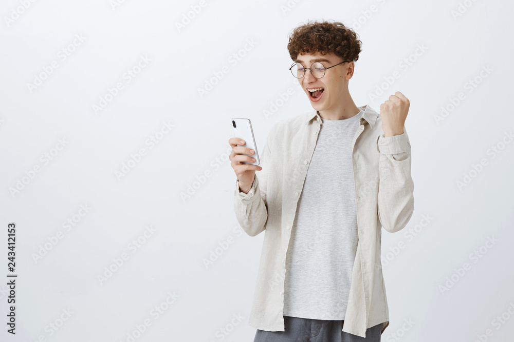 Portrait of pleased and triumphing stylish hipset gay man with curly hairstyle in round glasses clenching fist in victory and cheer gesture as smiling at smartphone screen, reading awesome news Portrait of pleased and triumphing stylish hipset gay man with curly hairstyle in round glasses clenching fist in victory and cheer gesture as smiling at smartphone screen, reading awesome news