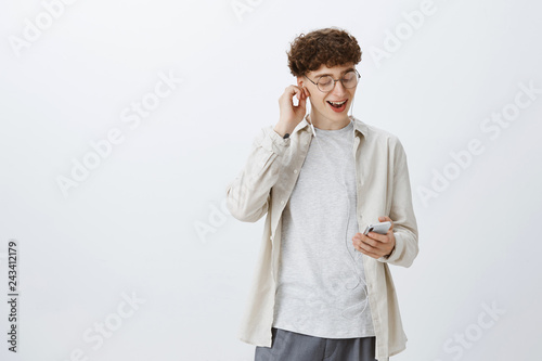 Excited and satisfied young good-looking hipster guy in round glasses with curly hair putting on earphone listening cool music in smartphone picking song as having good mood over gray background