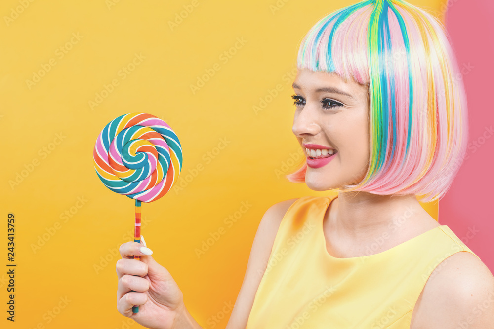 Woman in a colorful wig with a giant lollipop on a split yellow and pink background