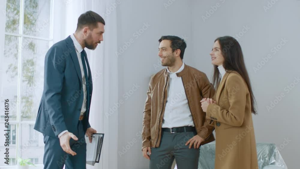 Professional Real Estate Agent Welcomes Young Couple at Home Showing. Young Couple Viewing New Property and Ready to Become Homeowners. Spacious Bright Apartment with Big Windows. Professional Real Estate Agent Welcomes Young Couple at Home Showing. Young Couple Viewing New Property and Ready to Become Homeowners. Spacious Bright Apartment with Big Windows.
